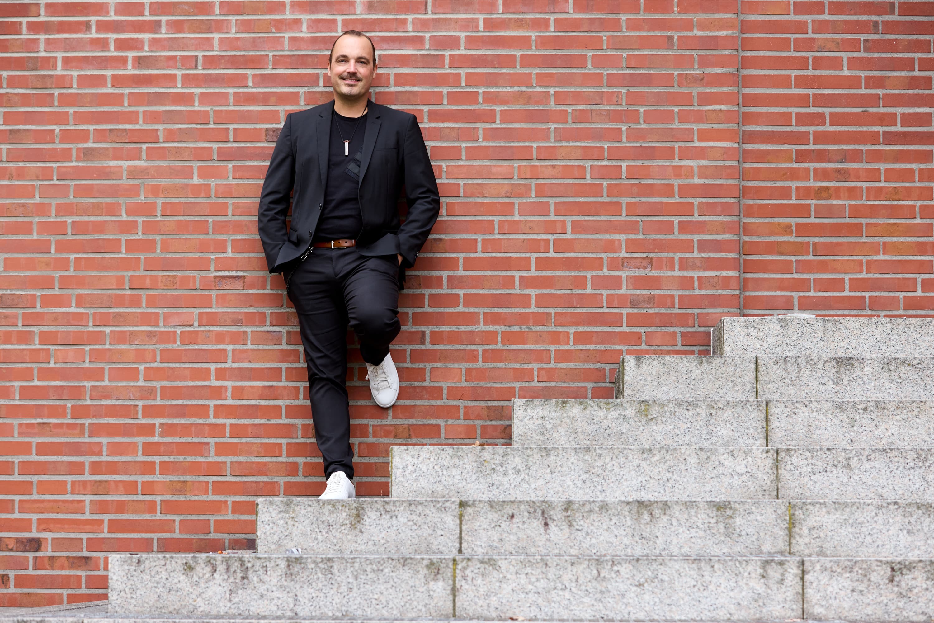 Markus Wessel lehnt lächelnd an einer Backsteinwand, weiße Sneaker, schwarzer Blazer